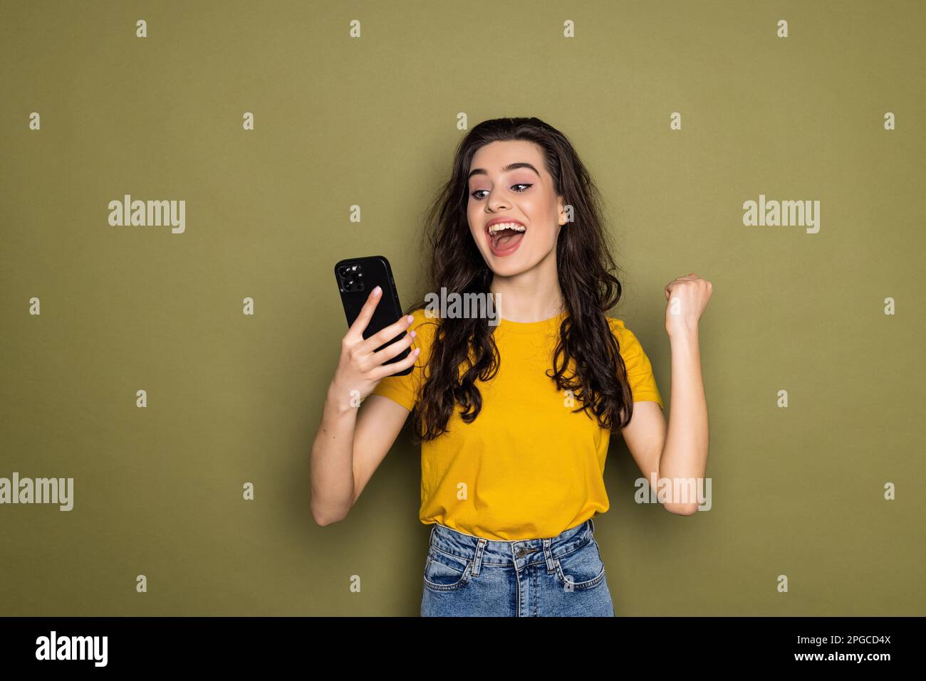 Frustrated shocked woman with long hair with smartphone in hands on khaki background looking at camera. Bad news, negative human emotions concept. Stock Photo