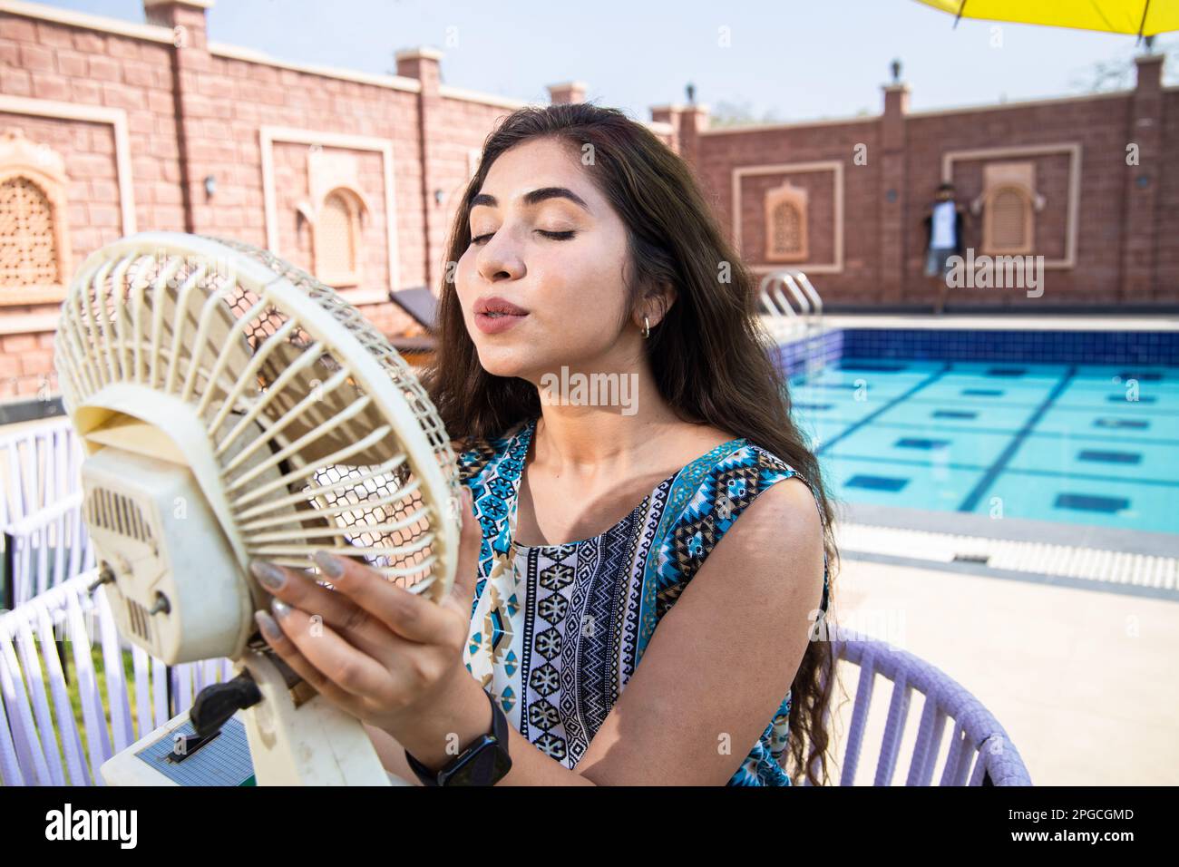 Young indian Woman enjoying air flow from electric fan and cooling her body outdoor in sunny day. Summer heat. Stock Photo