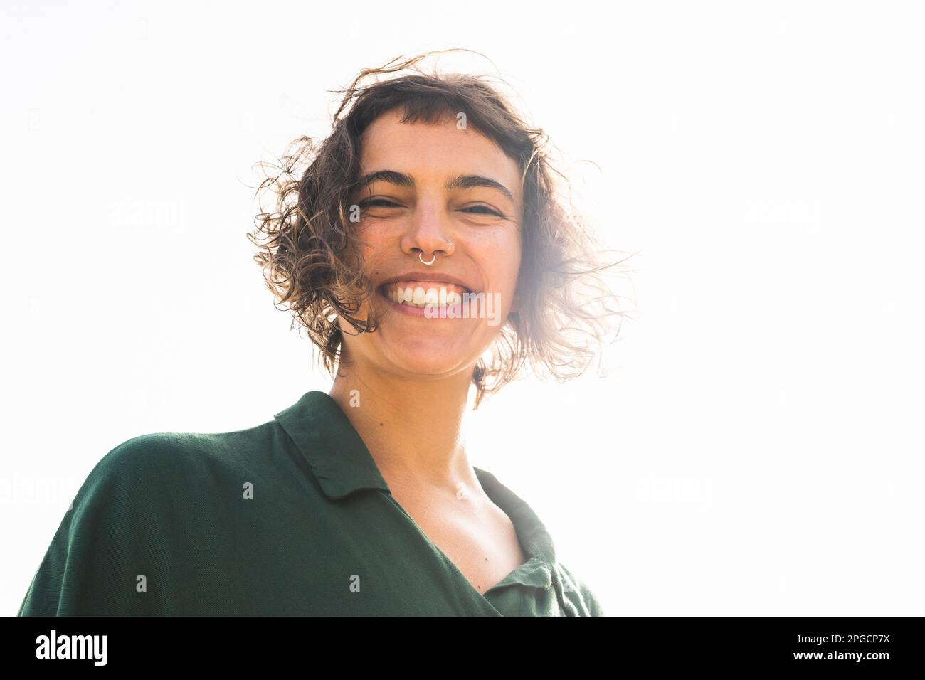 Low angle of happy young female with curly short hair and piercing smiling happily while looking at camera against bright light Stock Photo