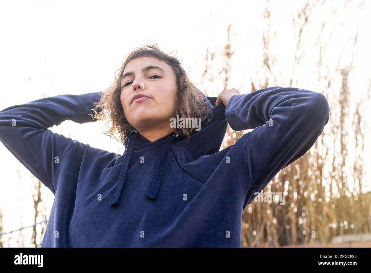 Low angle of calm young female with wavy hair and piercing wearing hoodie looking at camera on street Stock Photo