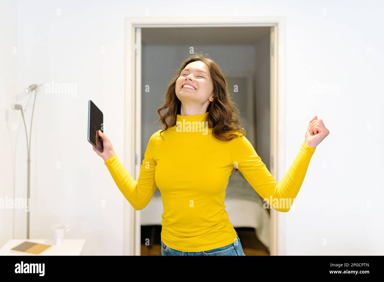 Delighted young female in yellow turtleneck smiling with eyes closed and holding tablet while standing celebrating victory with raised arm at home Stock Photo