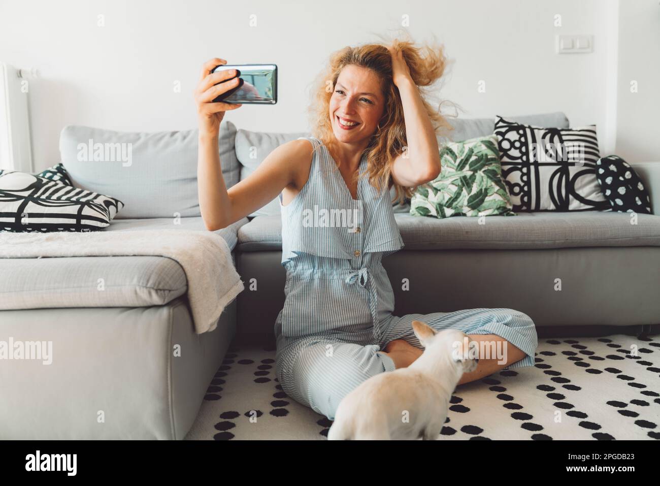 Smiling curly woman playing with her hair while taking selfies with her phone, sitting on the carpet by the couch Stock Photo