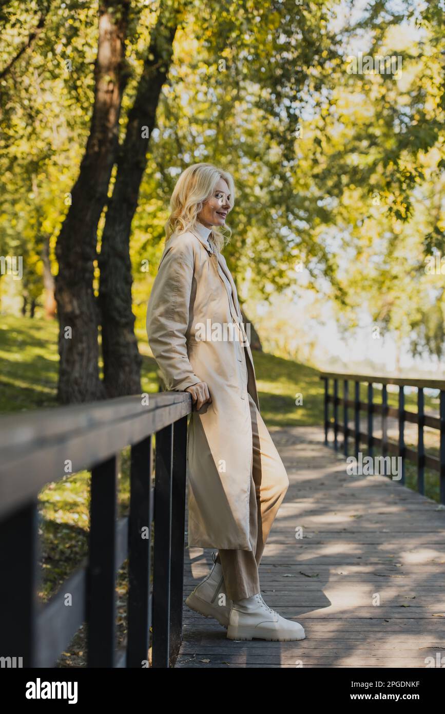 Mature blonde woman in trench coat smiling while standing on 