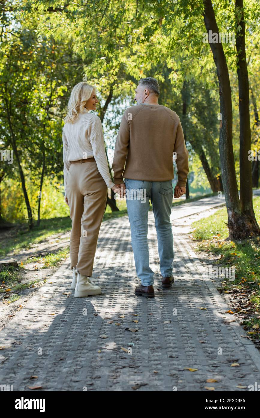 full length of stylish middle aged couple holding hands while walking together in green park,stock image Stock Photo