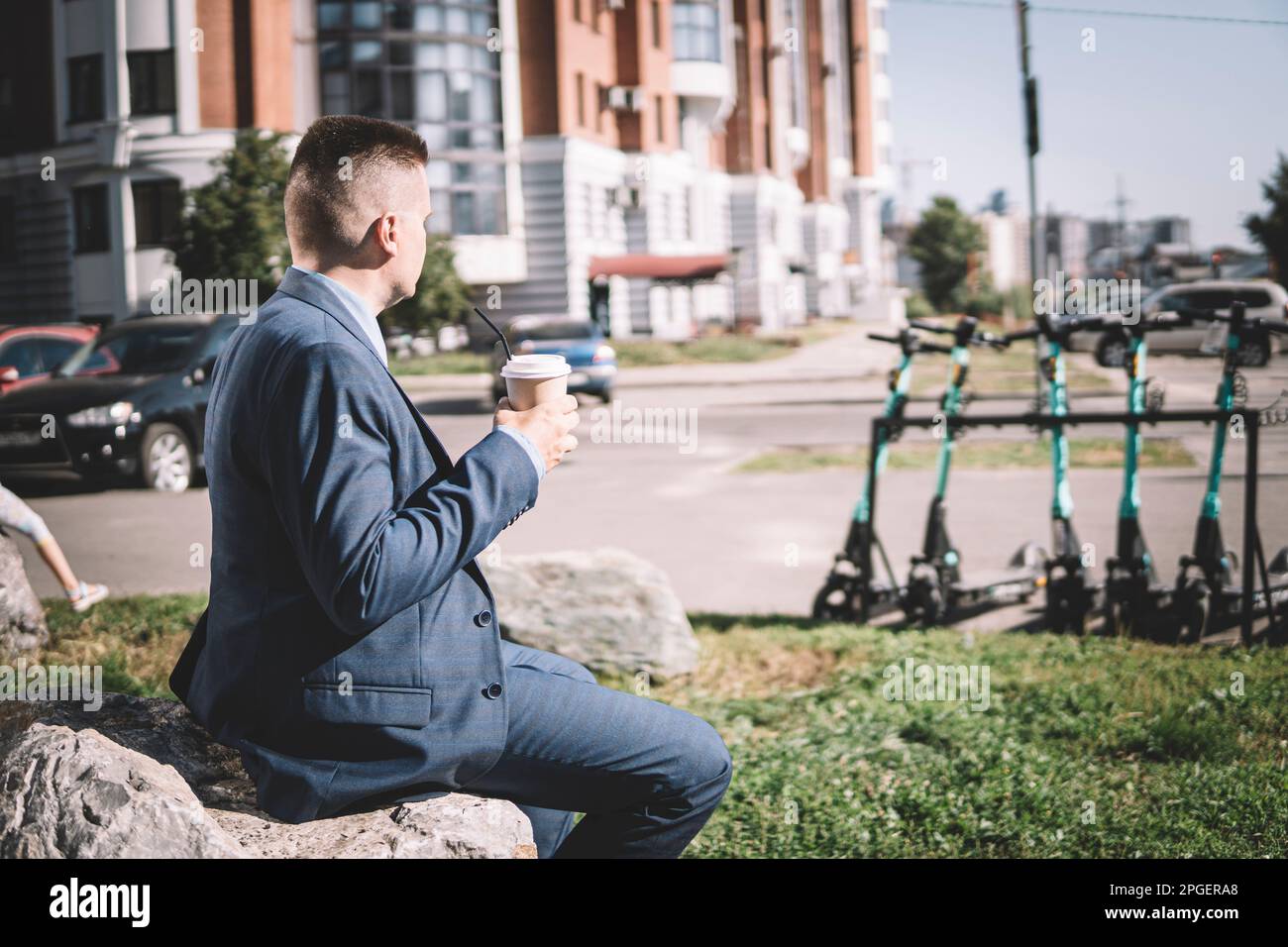 Young businessman in a business suit while look on electric scooter drinking coffee on the background of the morning city Stock Photo