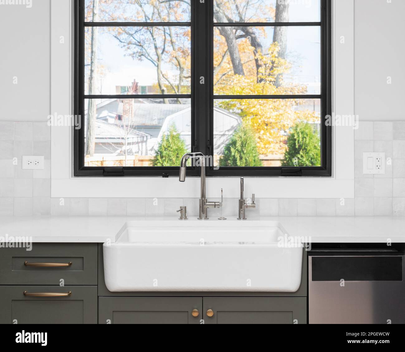 A sink detail shot with an apron sink, green cabinets, black window frame, and a tiled backsplash. Stock Photo