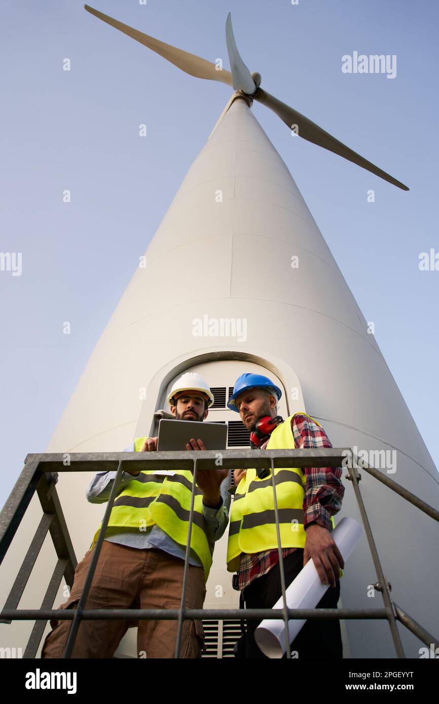 Vertical two helmeted engineers using and looking tablet. Professional technical at wind power plant Stock Photo