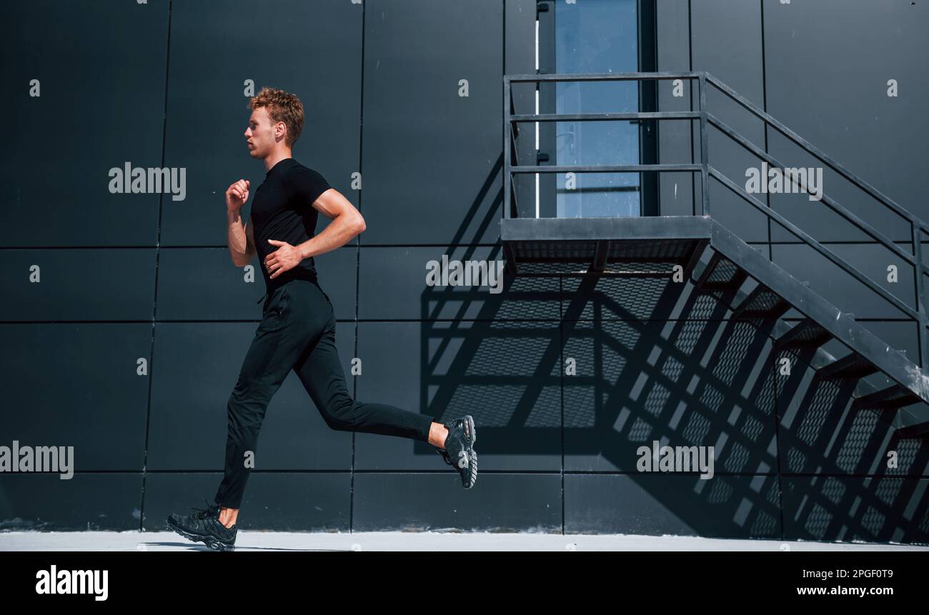 Running near black building. Sportive young guy in black shirt and