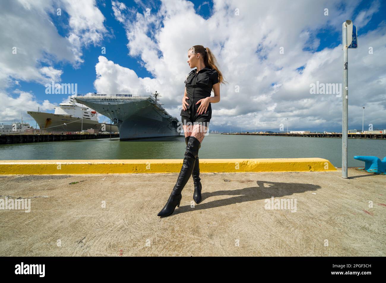 Fit Young Woman in Black Romper on Pier in Front of USS Aircraft Carrier | USS Hornet Stock Photo