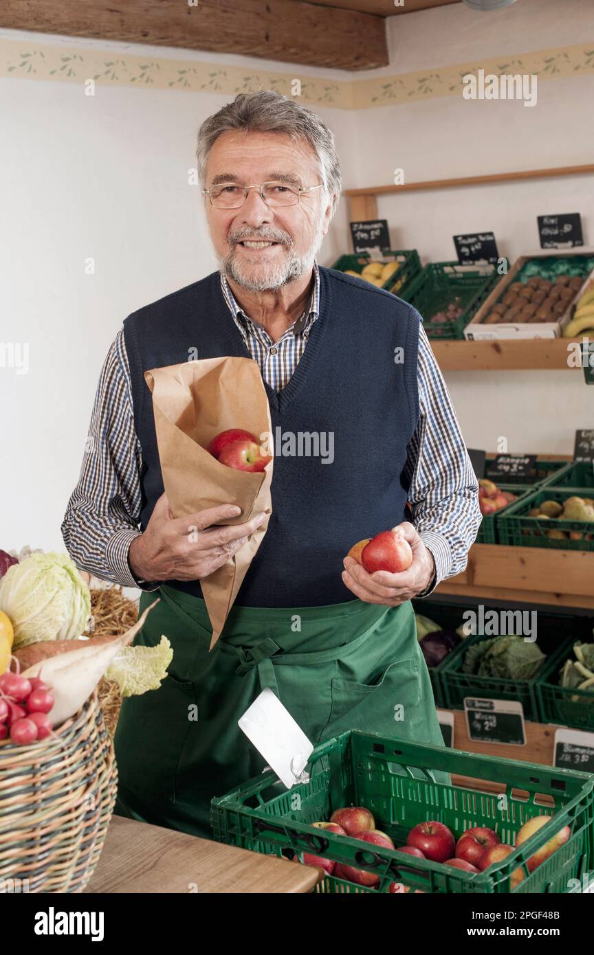 Portrait of a senior shopkeeper selling apples in  the store, Bavaria, Germany Stock Photo
