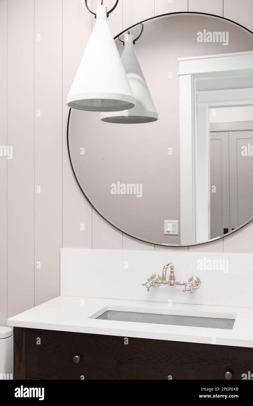 A bathroom detail with a dark wood cabinet, pendant light hanging down in front of a circular mirror, and a stone sink surrounded by a marble counter. Stock Photo