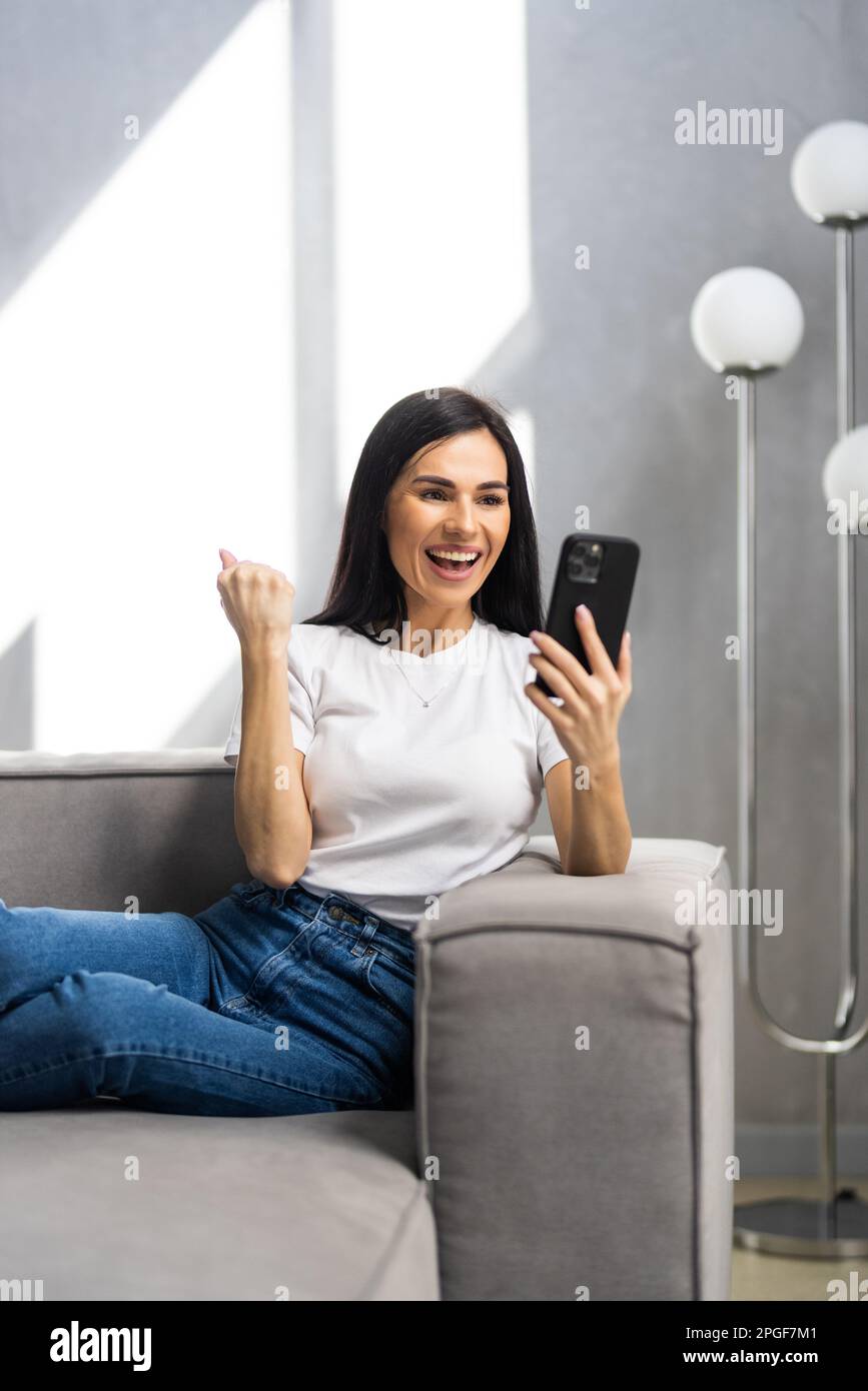 Excited woman use mobile phone raising fist up she celebrating victory win online or received good news on sofa living room at home. Happy female funn Stock Photo