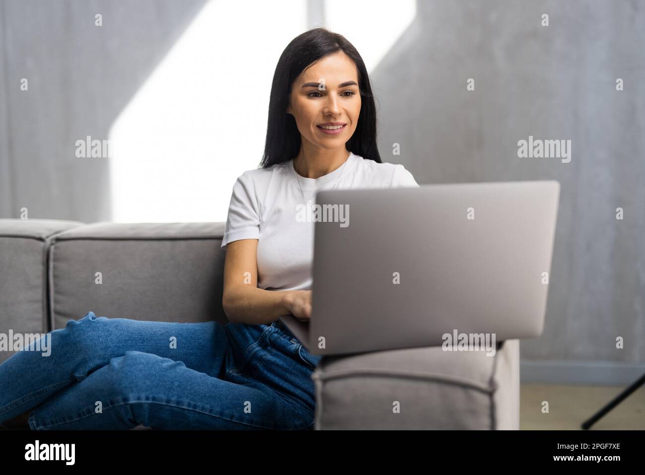 Photo of cheerful nice woman smiling and using laptop while sitting on couch in bright room Stock Photo