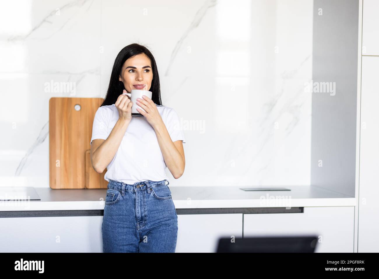 Positive caucasian woman drinking coffee and using digital tablet, surfing internet online, sitting at table in kitchen. Excited lady checking social Stock Photo