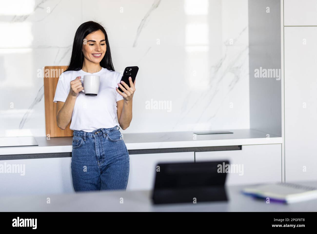 Young woman using smartphone leaning at kitchen table with coffee mug and organizer in a modern home. Smiling woman reading phone message. Brunette ha Stock Photo