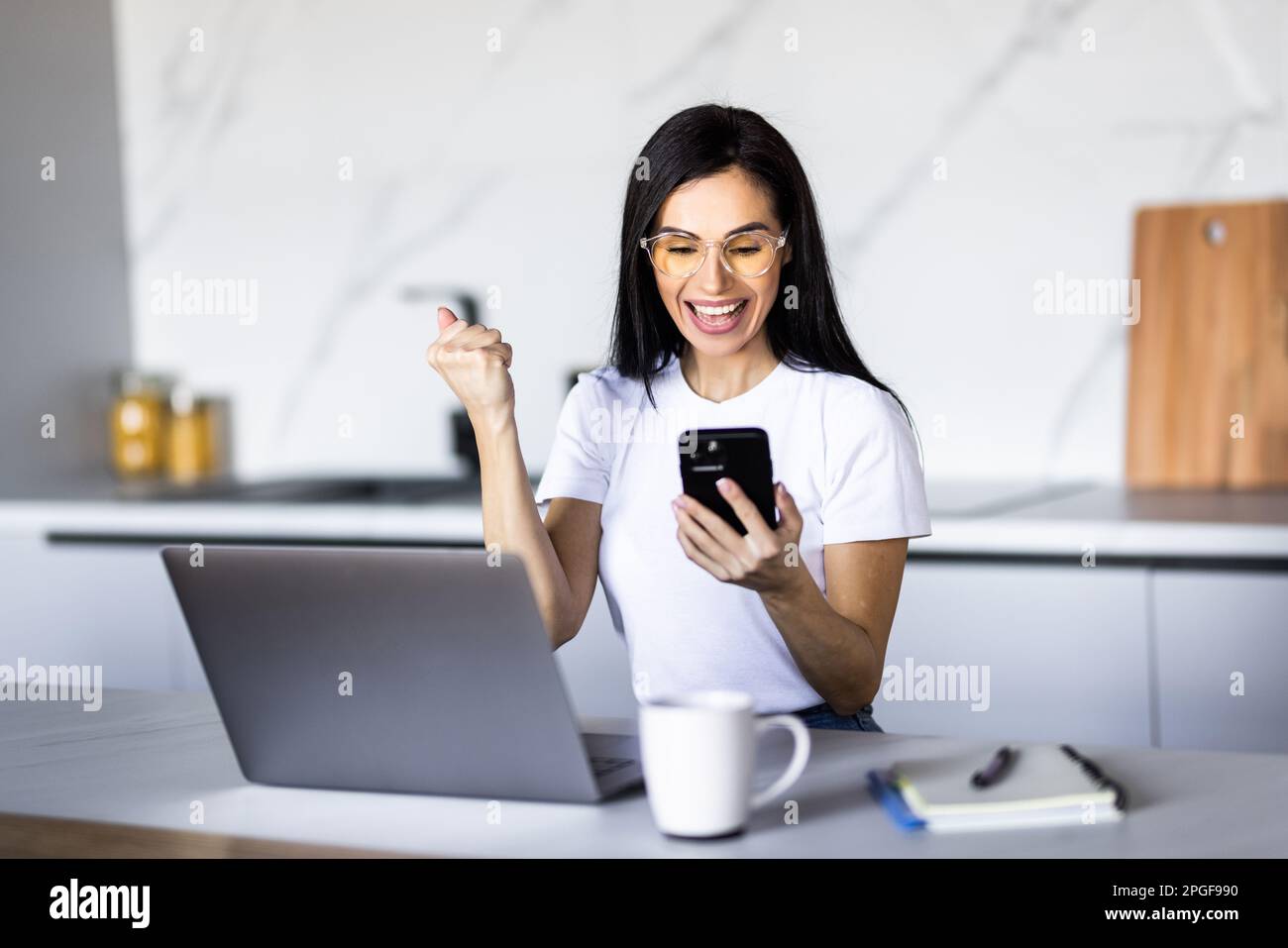Happy overjoyed woman with smartphone getting unbelievable good news from message on mobile phone while working at laptop from home, making yes winner Stock Photo