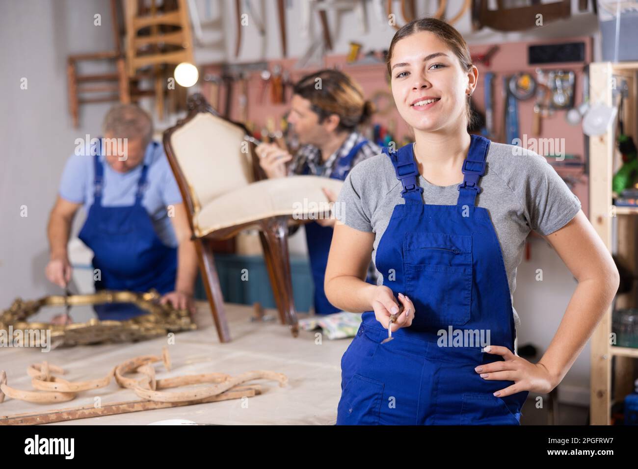Portrait of smiling skilled young European female furniture repair specialist in uniform posing in workshop Stock Photo