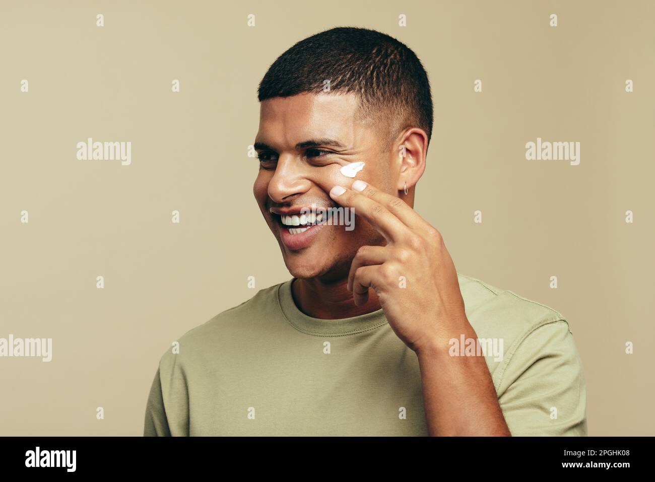 Youthful man smiles with joy as he introduces a beauty cream to his skincare routine. Happy young man pampering his face with a moisturizing product t Stock Photo