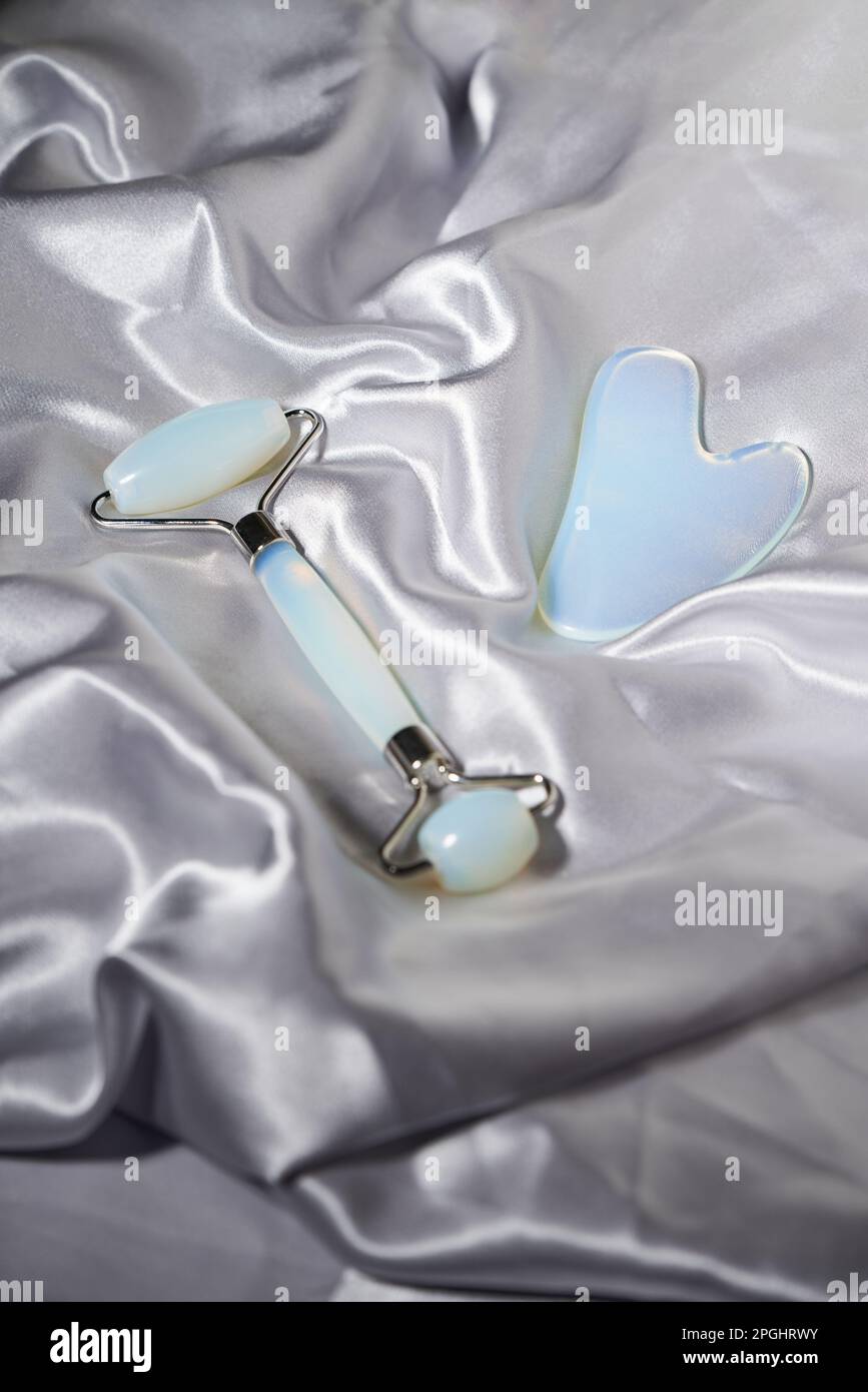 Blue jade face roller and Gua Sha scraping for beauty facial massage therapy on silver textured silk background. Face treatment and home spa, anti agi Stock Photo