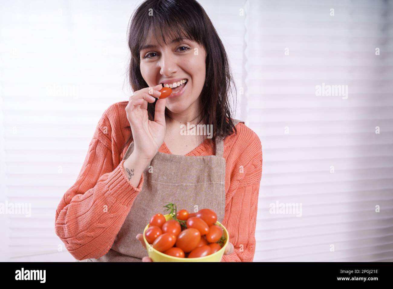 A Caucasian brunette housewife with long hair in an orange sweater and sand-colored apron bites a cherry tomato, holding a bowl by a backlit window, e Stock Photo