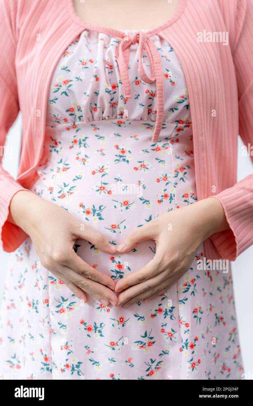 Prangnant woman in maternity dress putting her hands in heart shape over her belly standing against white backgrond, mother love concept Stock Photo