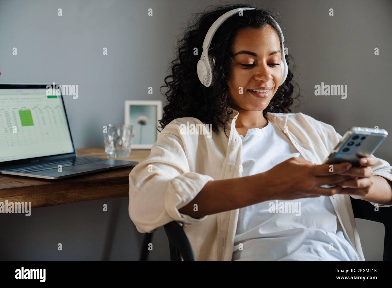 Black young woman in headphones using mobile phone while sitting on chair at home Stock Photo