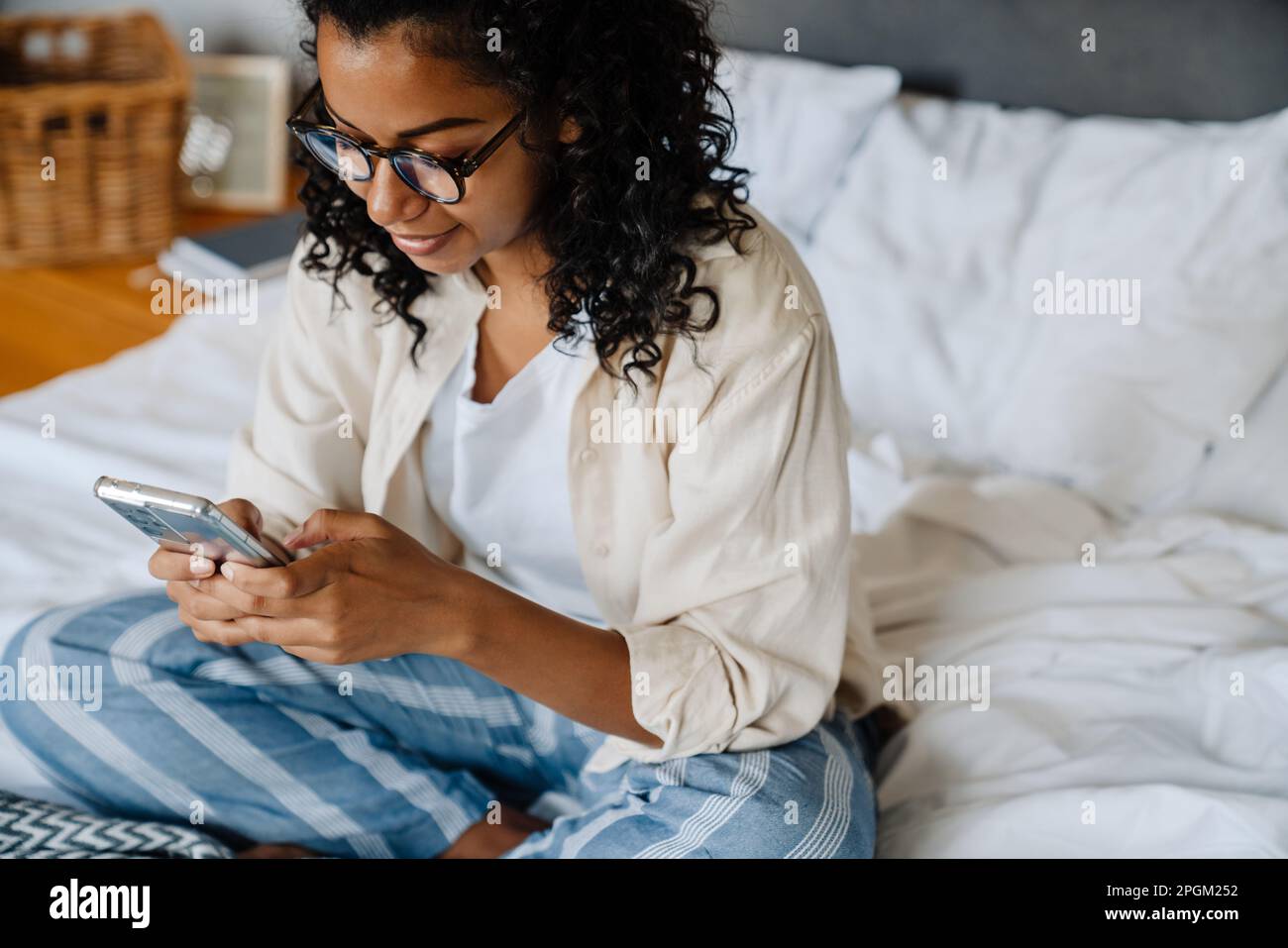 Black young woman in eyeglasses using mobile phone while resting on bed at home Stock Photo