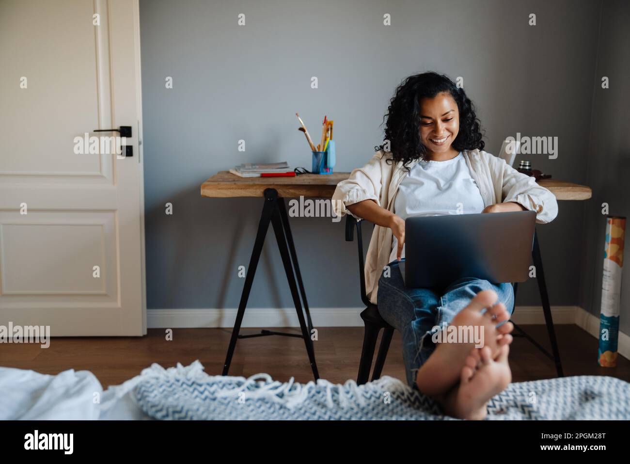 Black young woman smiling and working with laptop while sitting on chair at home Stock Photo