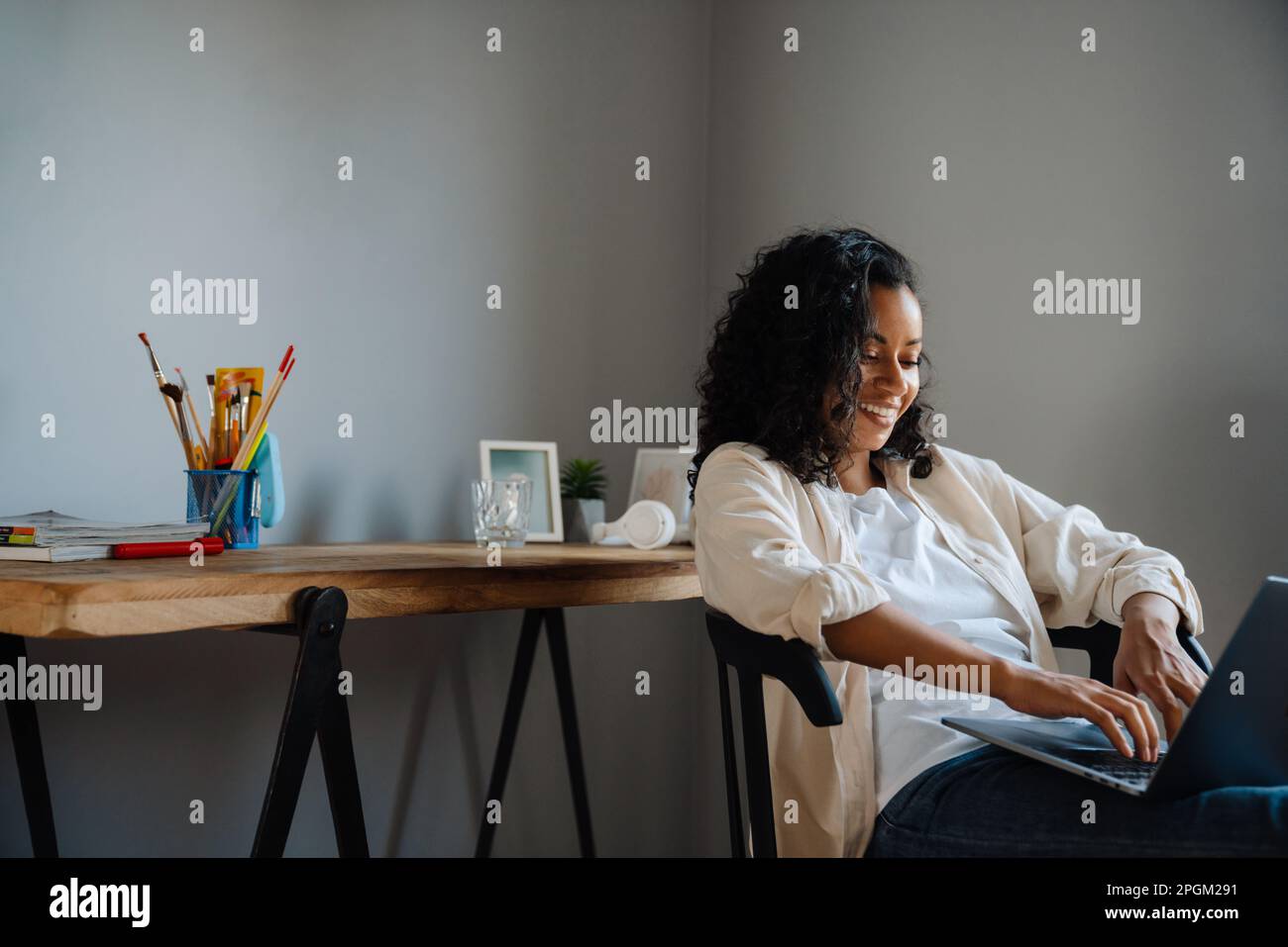 Black young woman smiling and working with laptop while sitting on chair at home Stock Photo