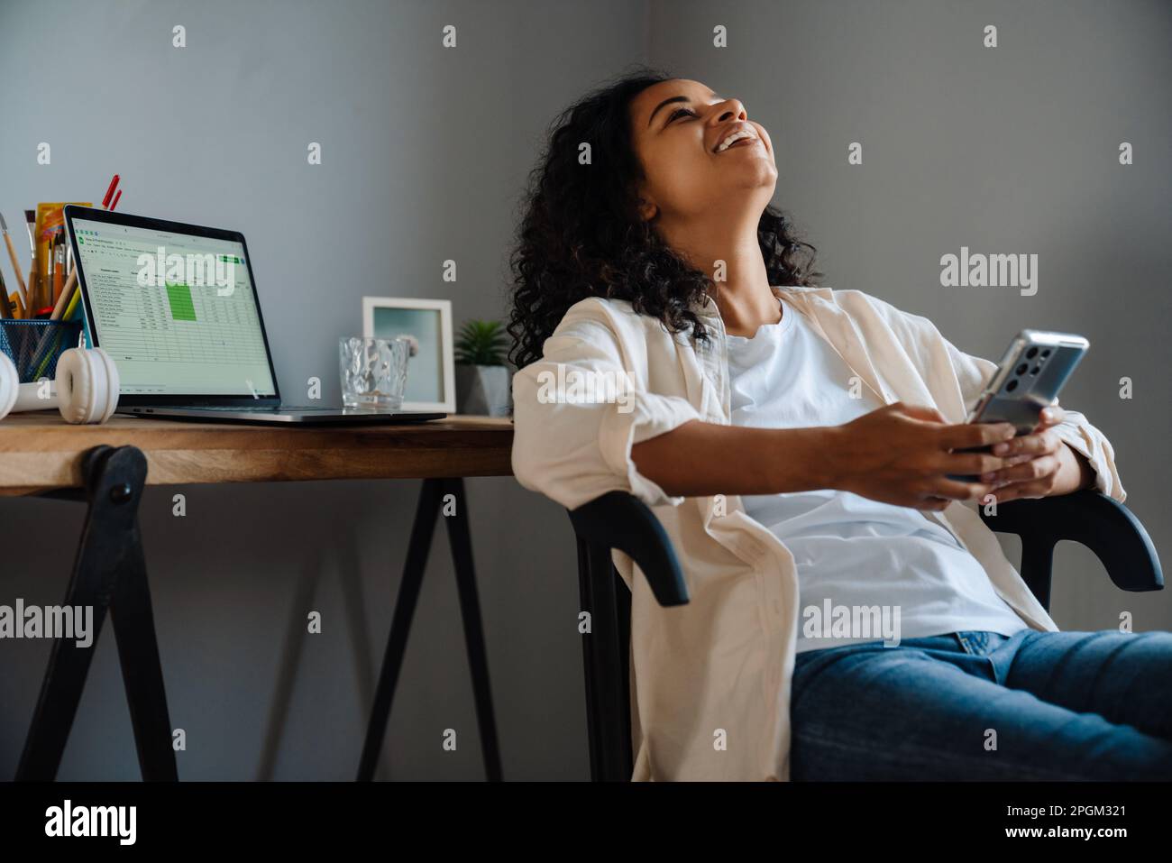 Black young woman smiling and using mobile phone while sitting on chair at home Stock Photo