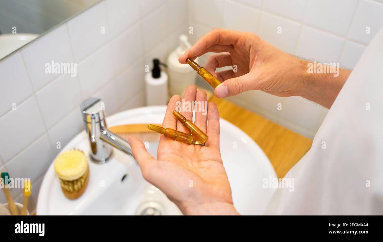 Male hands holding cosmetic ampoules with serum for hair growth, restoration beauty hair in a modern bright bathroom. Concept of hair loss and problem Stock Photo