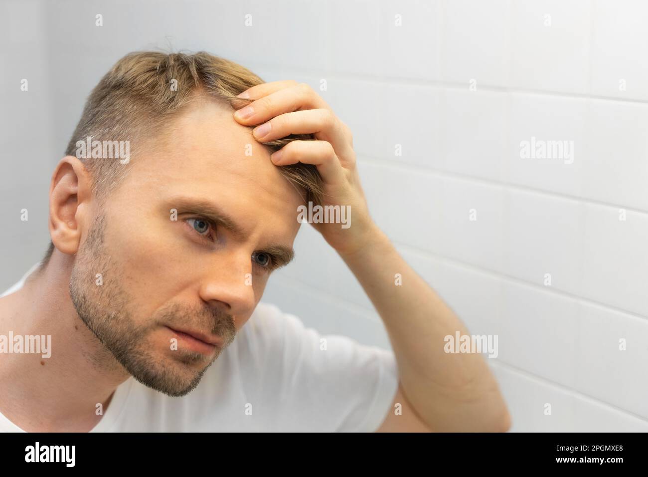 Middle aged caucasian white man with a short beard looks at his hair in the mirror in the bathroom and worried about balding. Male hair loss Stock Photo