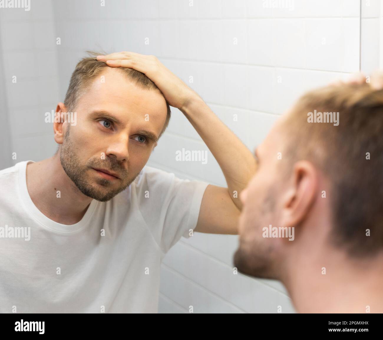 Middle aged caucasian white man with a short beard looks at his hair in the mirror in the bathroom and worried about balding. Man's hair loss Stock Photo
