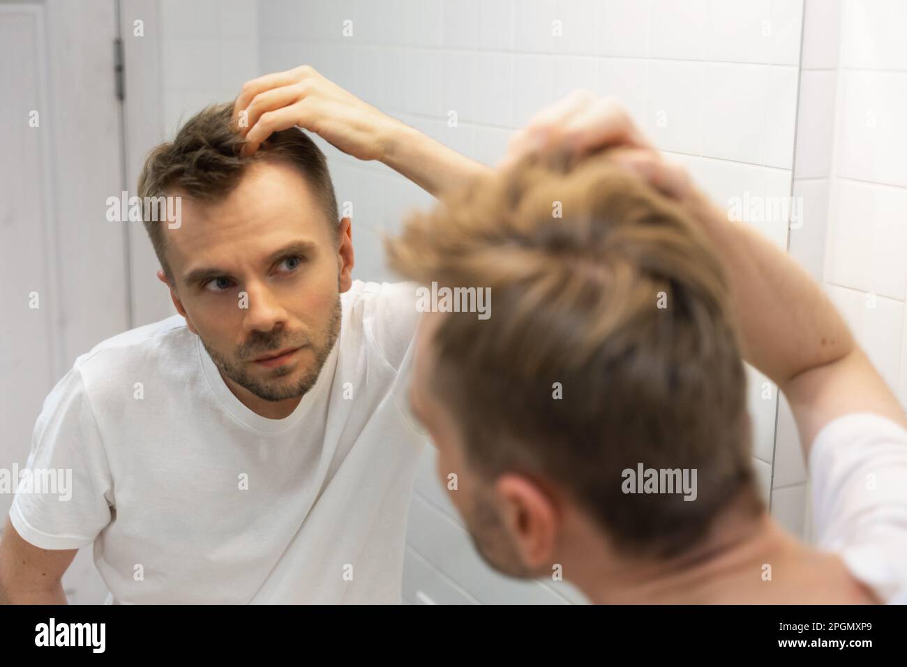 Middle aged caucasian white man with a short beard looks at his hair in the mirror in the bathroom and worried about balding. Man's hair problem Stock Photo