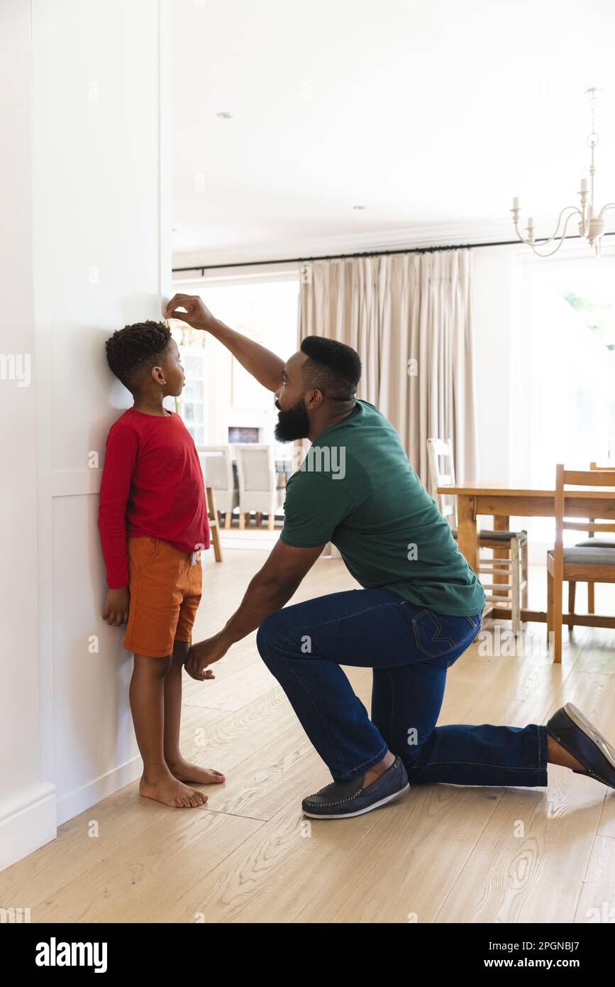 African american father and son measuring height against wall in kitchen. Family and domestic life. Stock Photo