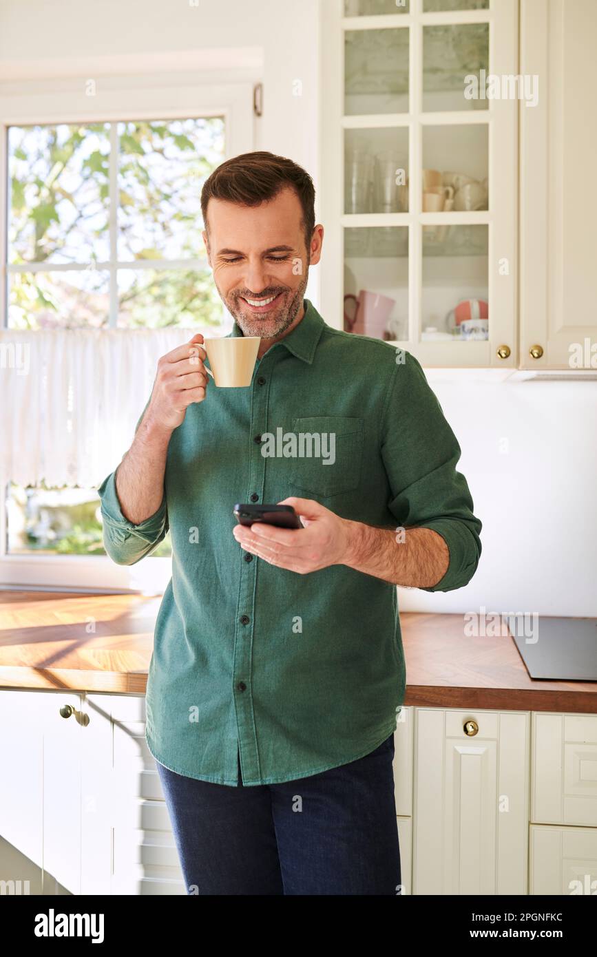 Smiling man using phone while drinking coffee in his kitchen Stock Photo