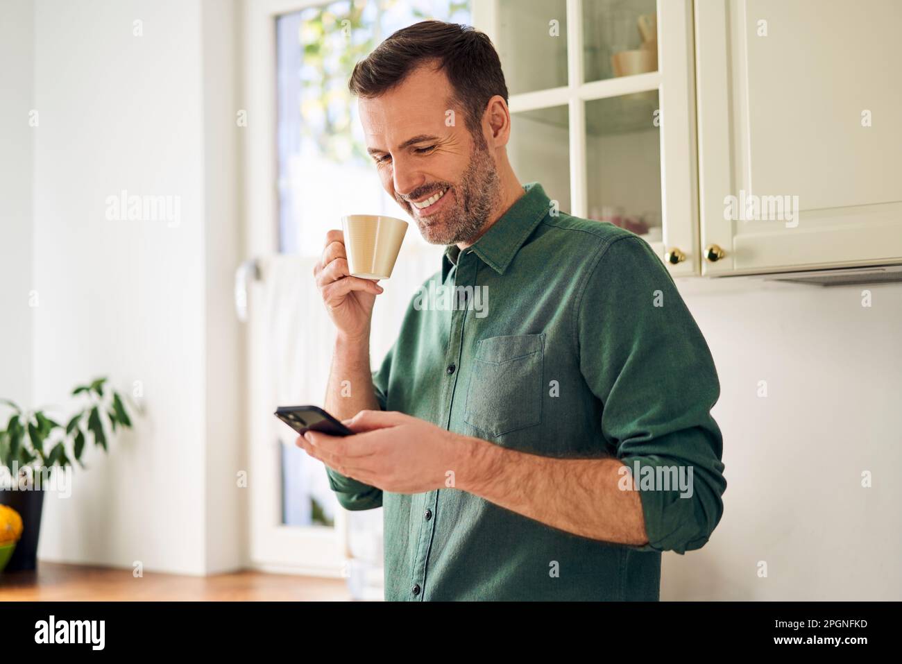 Smiling man using phone while drinking coffee in his kitchen Stock Photo