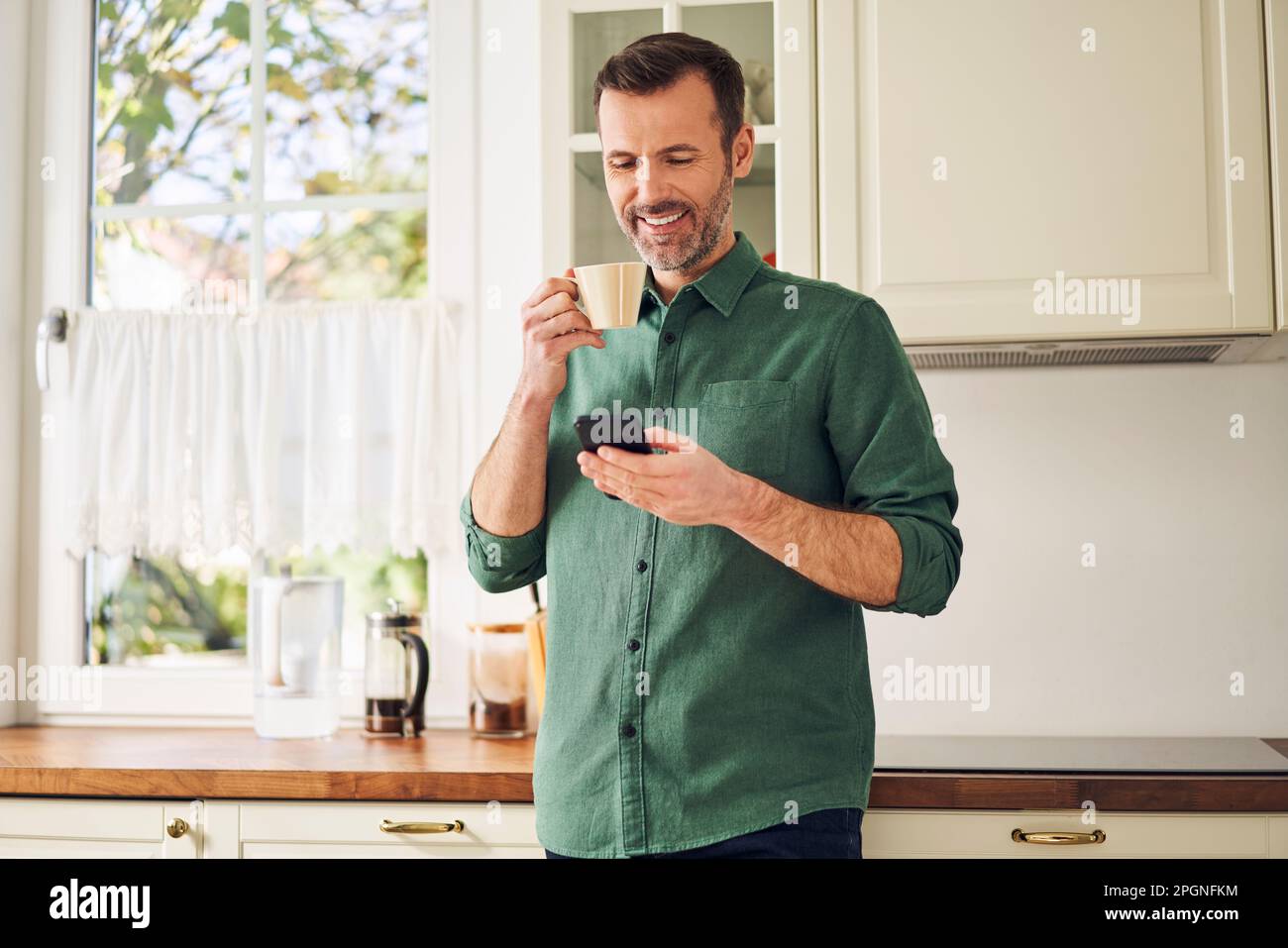 Smiling man using phone while drinking coffee in his kitchen Stock Photo