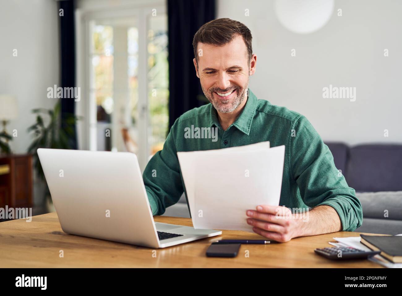Smiling man working from home using laptop holding documents Stock Photo