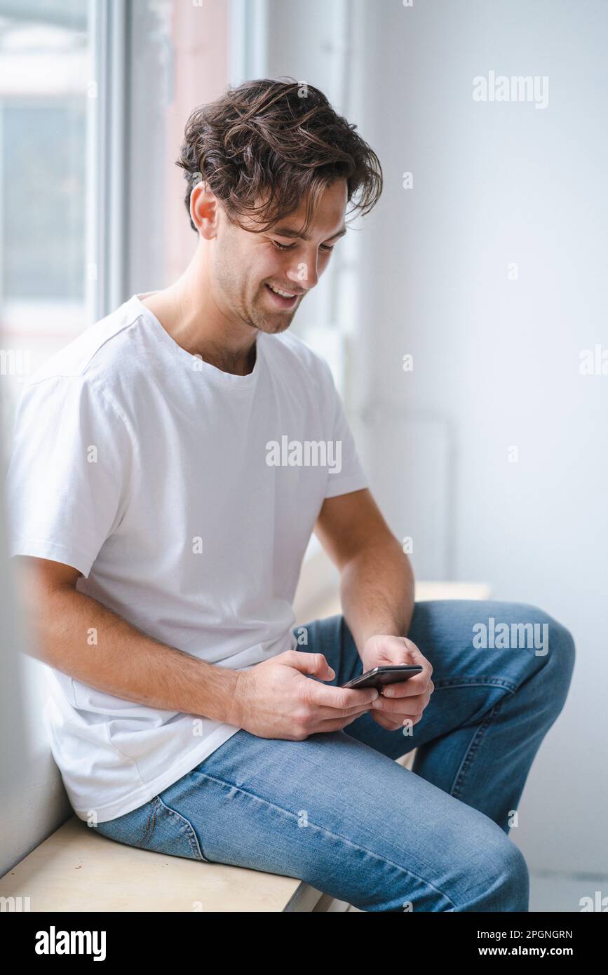 Happy young man using smart phone by window Stock Photo