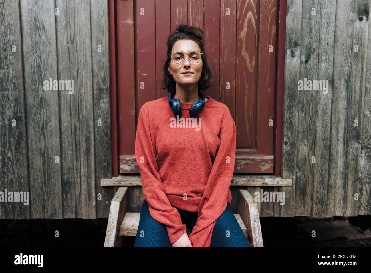 Smiling woman with wireless headphones sitting in front of door Stock Photo