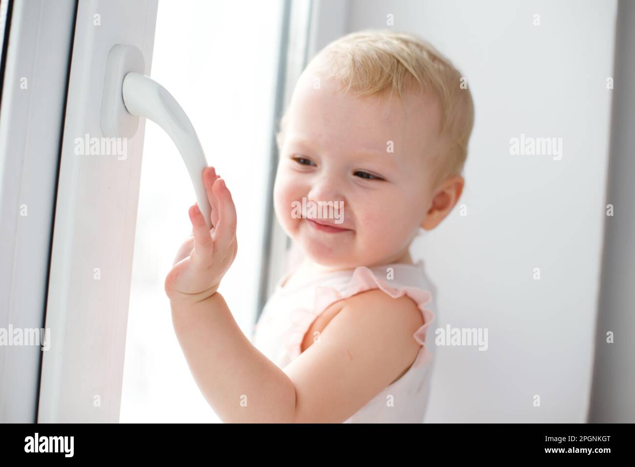 A little girl stands holding on to the handle of a broomtopplastic window. The danger of falling out of the window. The child stands on the windowsill Stock Photo