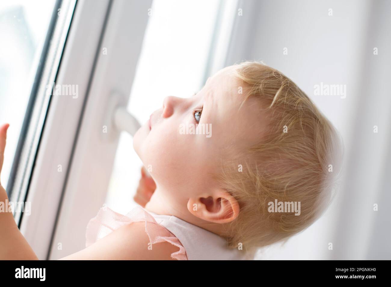 Protecting children from falling out of windows. An active little girl carefully opens a metal-plastic window in the house. Stock Photo