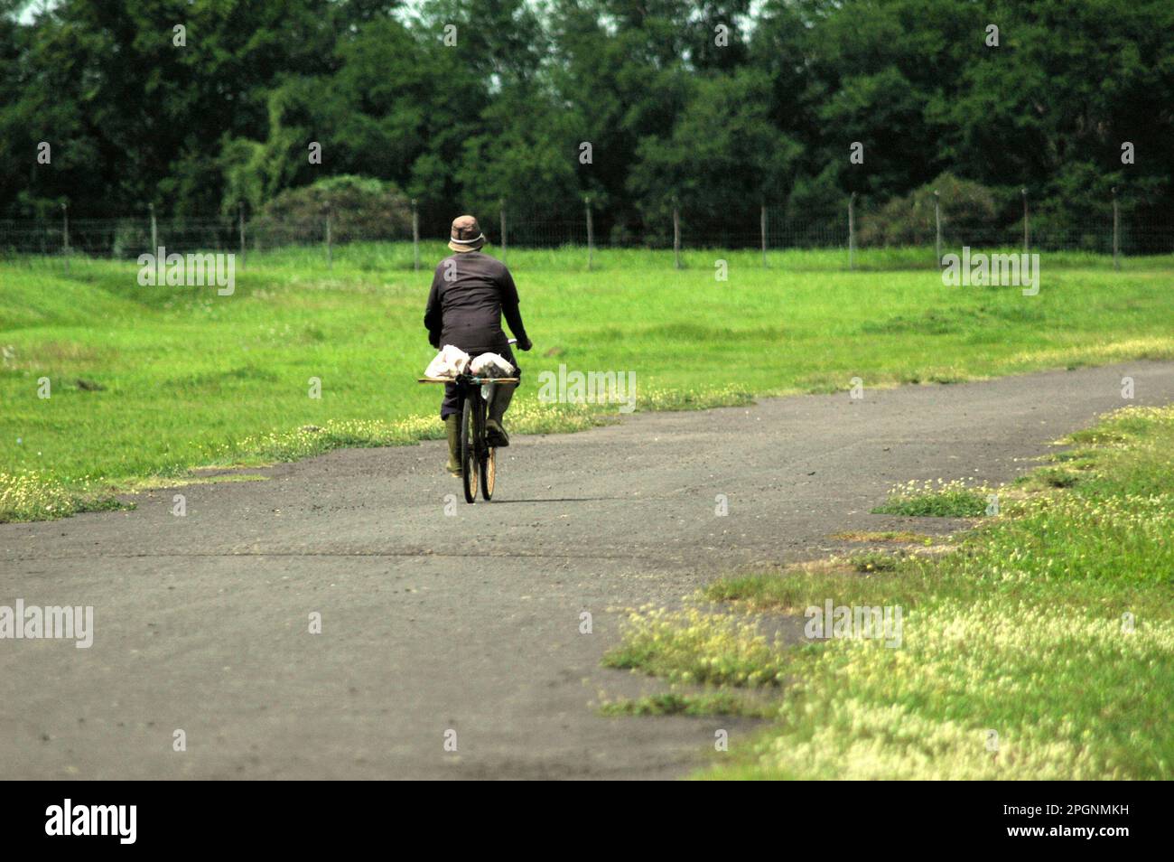 A man is riding his bicycle on a road in the buffer zone of Jakarta International Soekarno-Hatta Airport in Rawa Bokor, Tangerang, Banten, Indonesia. The buffer zone of Jakarta's Soekarno-Hatta International Airport, which consists of wetland and grassland areas neighboring the airport and human settlements, is somehow offering beautiful views. It serves ecologically as a water catchment area. As the grassland allows local community to let their goats grazing, wildlife—dominated by various waterbird species—enjoys the wetland as a transit and feeding ground. Stock Photo