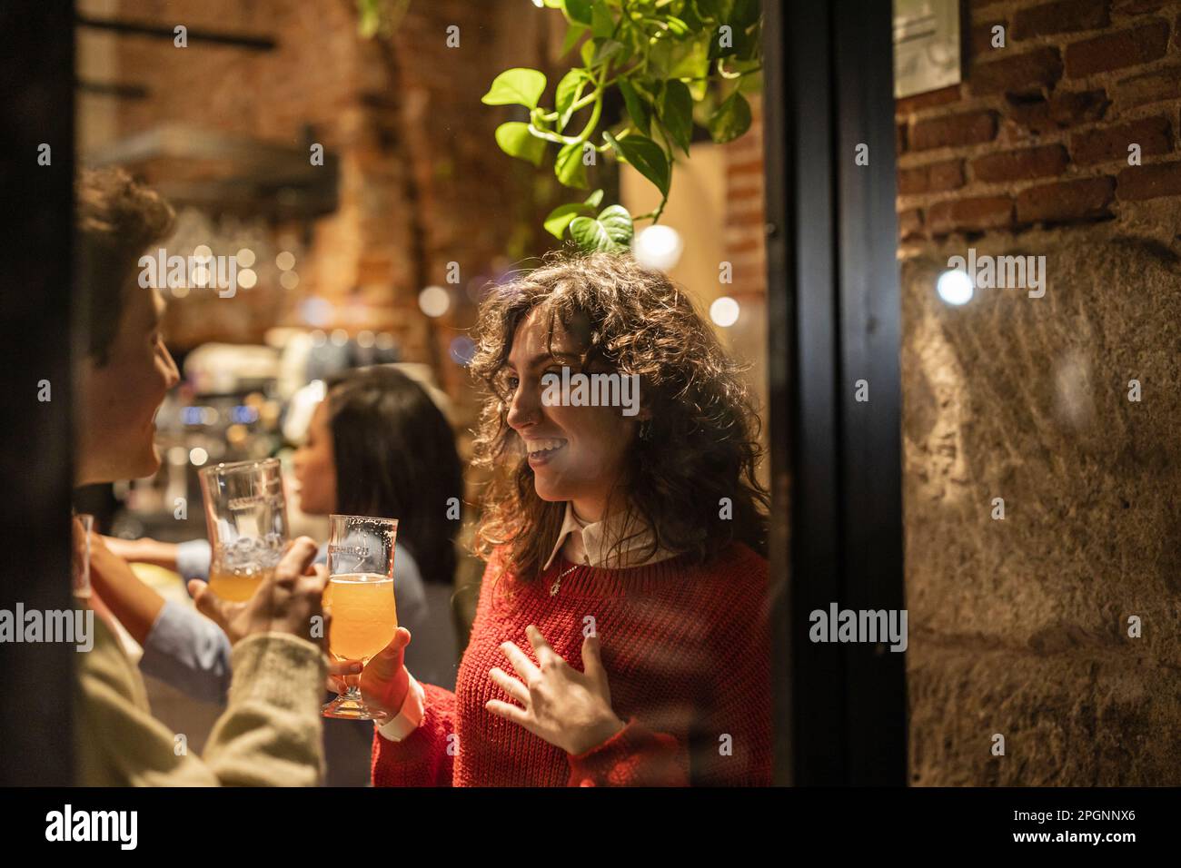 Young woman with glass of beer standing with friend seen through window Stock Photo