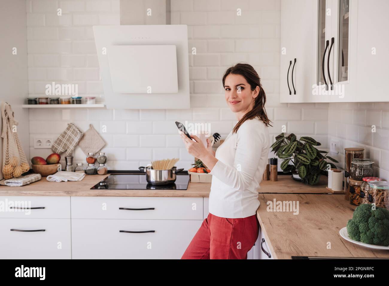 Smiling woman with mobile phone standing by counter in kitchen Stock Photo