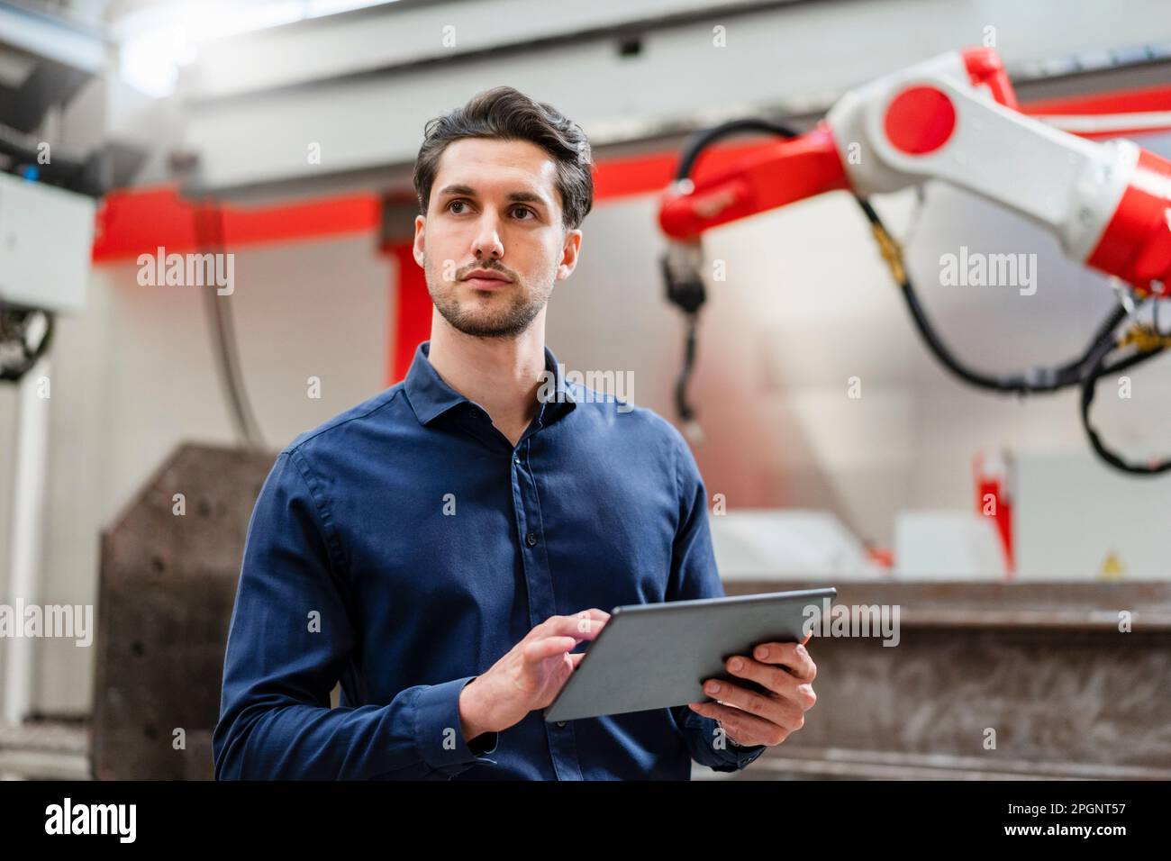 Thoughtful engineer holding tablet PC standing in robot factory Stock Photo