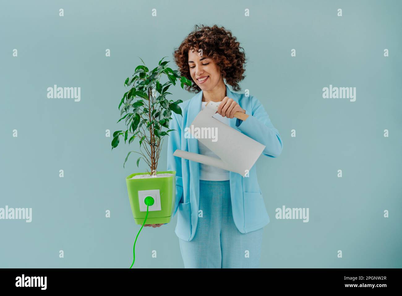 Happy woman watering potted plant connected with electric plug against blue background Stock Photo