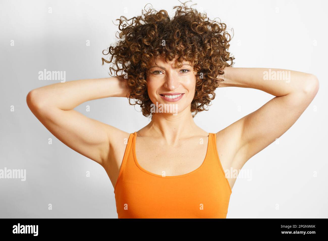 Smiling curly haired woman standing with hands behind hair against white background Stock Photo