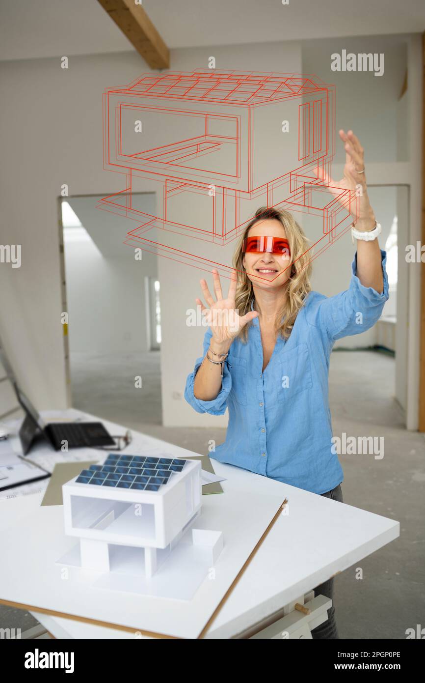 Smiling mature architect wearing virtual reality simulator gesturing and examining house model Stock Photo